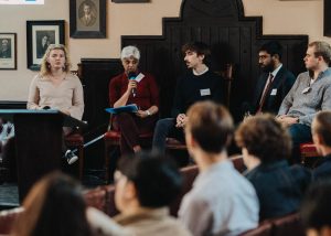 A panel discussion in a formal setting. Five individuals are seated on a stage, with one woman in a red top holding a microphone and speaking. The others appear to be listening attentively. The audience is partially visible in the foreground, blurred. The room is adorned with framed portraits on the walls, and the seating consists of leather chairs. The atmosphere suggests an academic or professional event.