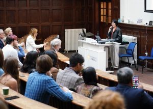 A speaker stands at a lectern in a traditional lecture hall with wood-paneled walls, addressing an audience seated in tiered rows. The audience consists of individuals attentively listening, some holding notebooks or phones. The lectern is branded with the University of Cambridge logo, and there is a projection screen visible on the right.
