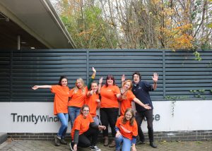 A group of eight cheerful volunteers posing in front of a sign that reads "Trinity Winchester." Seven of them are wearing bright orange T-shirts with "Volunteer" written on them, while one person is dressed in a navy blue top. They are smiling, waving, and striking playful poses, with trees and a modern fence visible in the background.