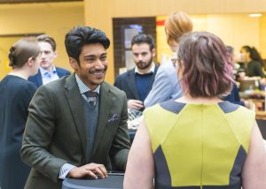 A group of professionally dressed individuals engaged in conversation at a networking event. The foreground features a man in a dark green suit with a pocket square speaking to a woman wearing a green and black dress. The background shows other attendees mingling in a brightly lit venue with refreshments visible on tables.