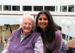 Two individuals are pictured indoors, seated closely together. The person on the left is an older adult with white hair, wearing a purple knitted vest over a floral blouse. The person on the right has long dark hair and is wearing a purple top and jeans, leaning slightly toward the older adult. They are in a bright, spacious setting with large windows and modern decor in the background, including a mobility aid and wicker chairs.