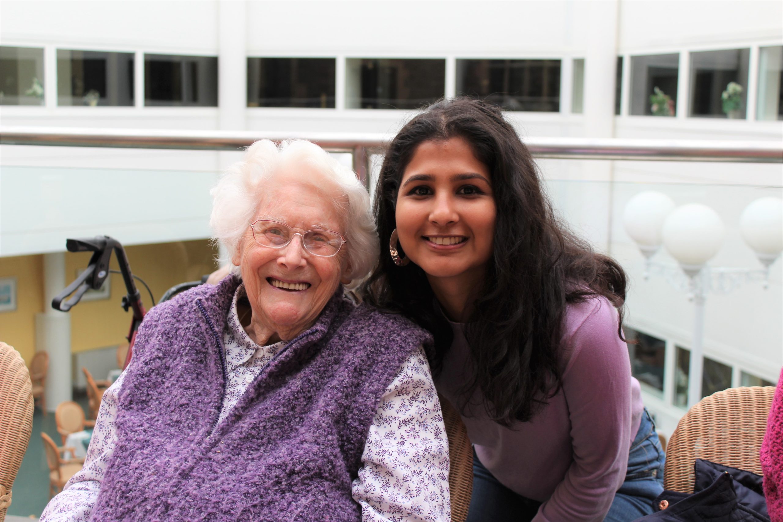 Two individuals are pictured indoors, seated closely together. The person on the left is an older adult with white hair, wearing a purple knitted vest over a floral blouse. The person on the right has long dark hair and is wearing a purple top and jeans, leaning slightly toward the older adult. They are in a bright, spacious setting with large windows and modern decor in the background, including a mobility aid and wicker chairs.