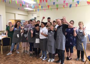 A group of people standing together in a brightly decorated community hall with colorful bunting hanging from the ceiling. Many are wearing matching grey aprons with "The Community Café" logo, smiling and waving at the camera. The room has wooden flooring, chairs, and tables in the background, creating a welcoming and lively atmosphere.