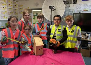 A group of six children and one adult are posing in a classroom setting. They are wearing high-visibility vests and safety goggles, and some are holding tools such as a tape measure and a drill. In front of them on a pink tablecloth is a wooden birdhouse, along with screws and other small tools. The background features educational posters, a large light reflector, and a screen. The group appears to be engaged in a hands-on woodworking or crafting activity.
