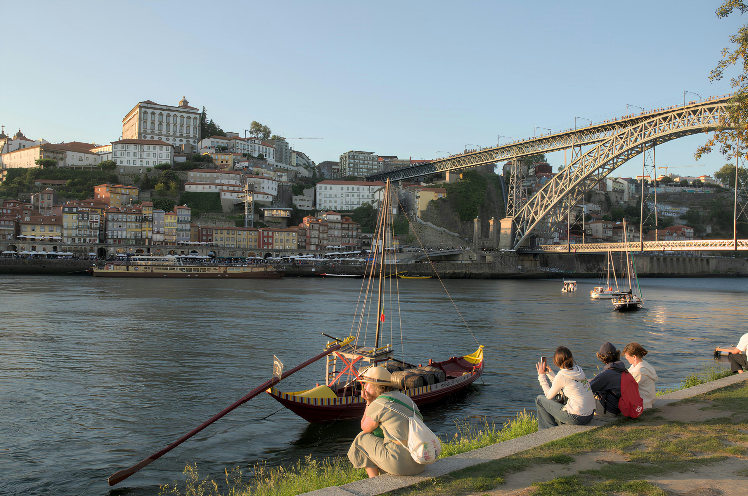 A riverside scene in Porto, Portugal, showing people sitting on the grassy bank of the Douro River. In the foreground, a traditional Rabelo boat with barrels is moored on the water. Across the river, colorful buildings rise up the hillside beneath the prominent Dom Luís I Bridge, an iconic double-deck metal arch bridge. The cityscape is bathed in warm evening light, and the sky is clear. Photo by Tayan Lima on Unsplash
