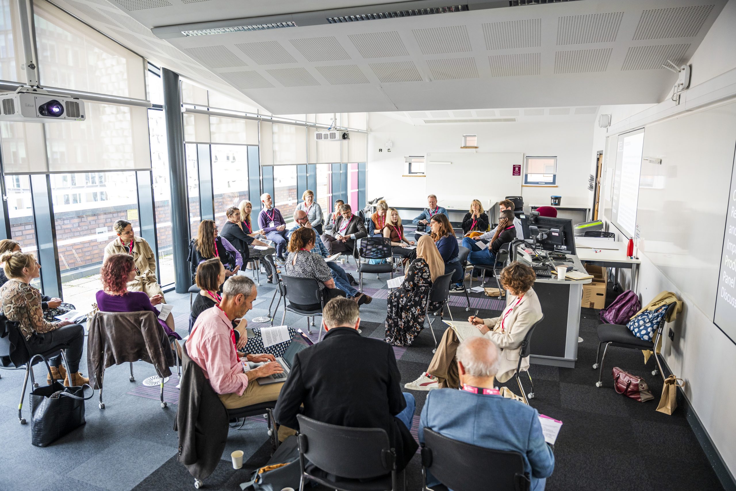 Conference participants seated in a large circle inside a modern, well-lit room with floor-to-ceiling windows. Attendees are engaged in discussion, taking notes, and wearing event lanyards. The setting features chairs, tables, and presentation equipment, suggesting an interactive workshop or group session. Coats and bags are placed around the room, contributing to a casual, collaborative atmosphere.