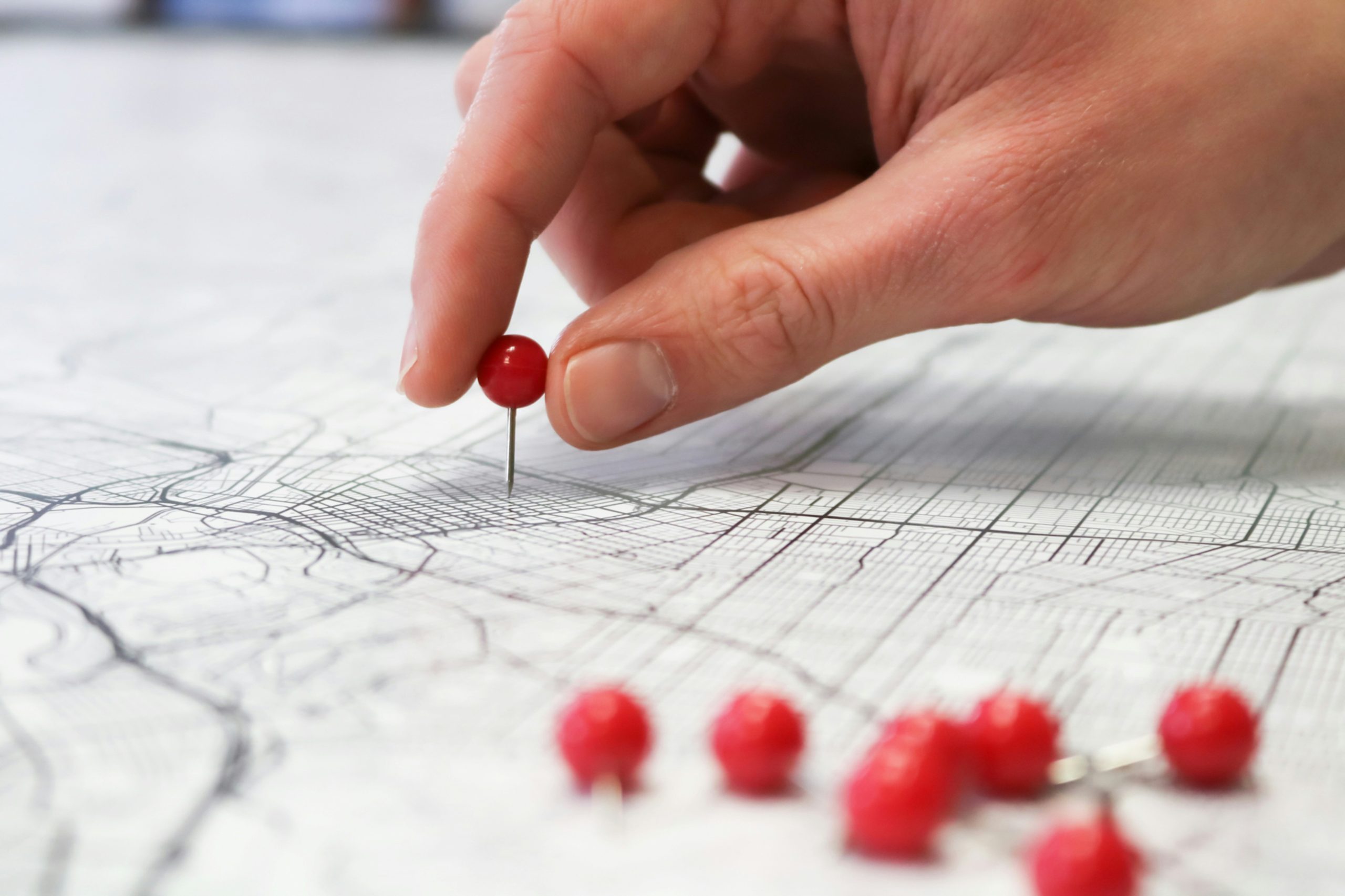 A close-up of a hand placing a red pushpin into a map, with several other red pushpins lying on the map, representing the marking of specific locations or points of interest.