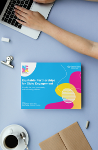 A person's hands are typing on a laptop on a pastel blue desk. Below the laptop is a brightly coloured booklet titled 