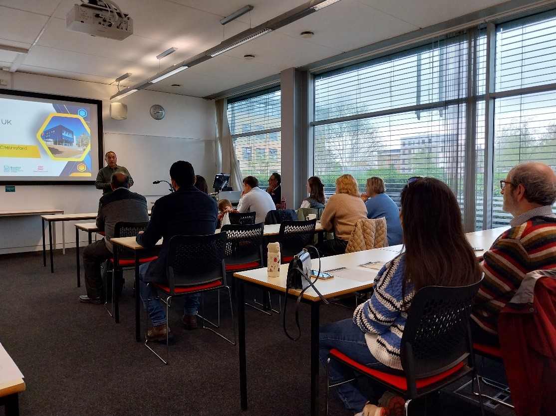 People attending a classroom presentation, seated at tables and listening to a speaker at the front with a projected slide visible on the screen.