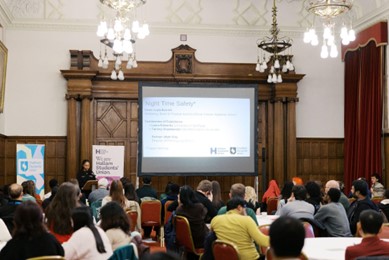 People seated at round tables in a large hall listen to a speaker presenting a slide on a projector screen at the front of the room.