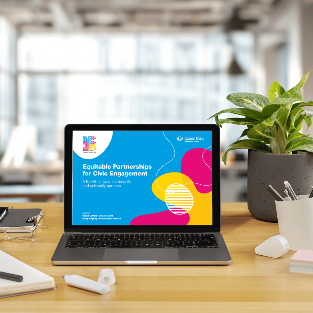 A modern workspace with a wooden desk holding an open laptop displaying a colorful presentation slide titled "Equitable Partnerships for Civic Engagement" by Queen Mary University of London and the National Civic Impact Accelerator. The slide includes author names and bright abstract shapes in blue, yellow, and pink. Surrounding the laptop are stationery items including papers, glasses, a plant in a grey pot, a white cup with pens, and a white marker pen cap. The background features a bright, softly blurred office environment with large windows.