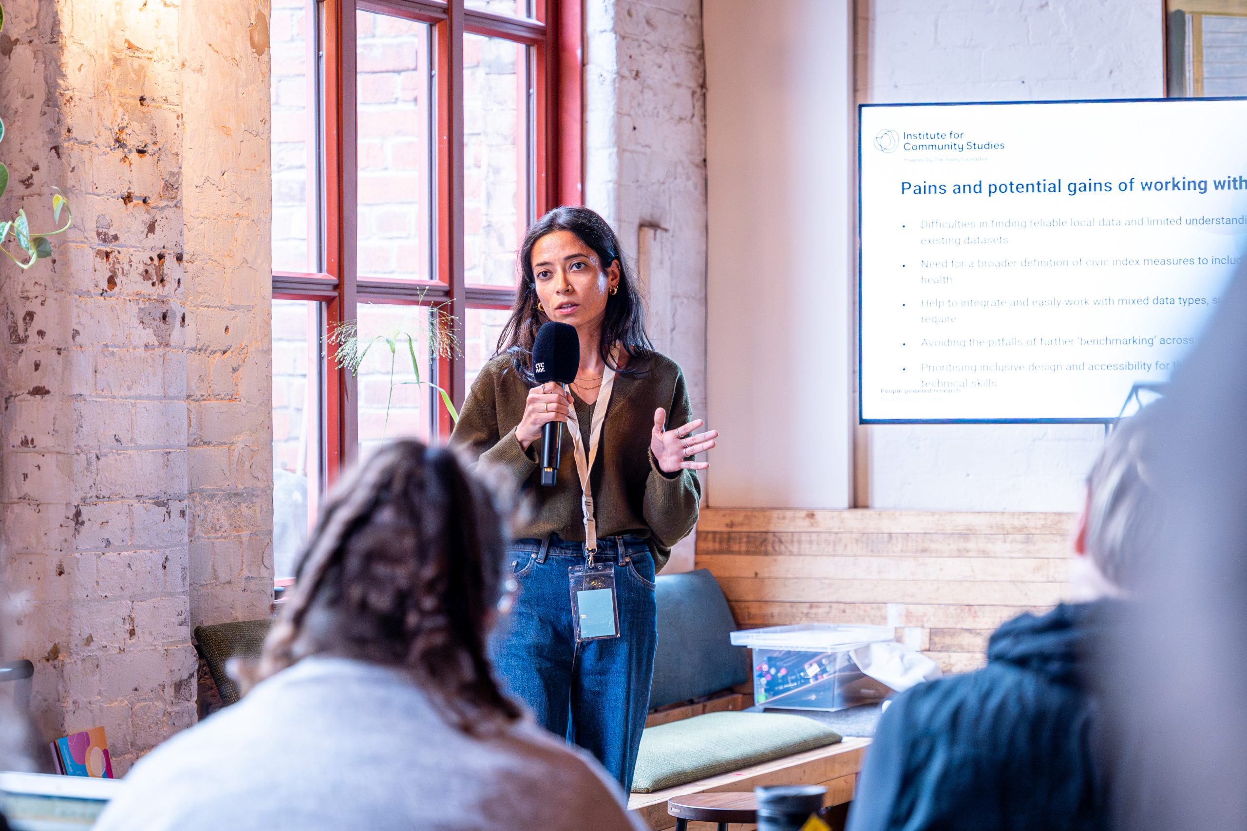 Person giving a presentation with a microphone in a casual indoor setting, speaking to an audience with a projected slide visible in the background.